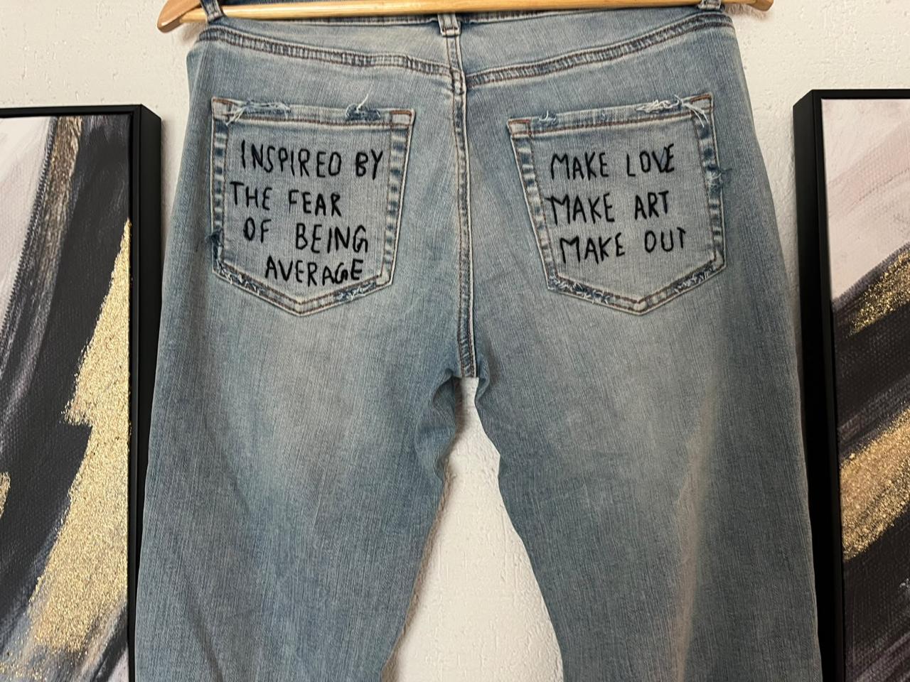 Back of: Light blue flared jeans, embroidered back pockets: "Inspired by the fear of being average" and "Make love, make art, make out"