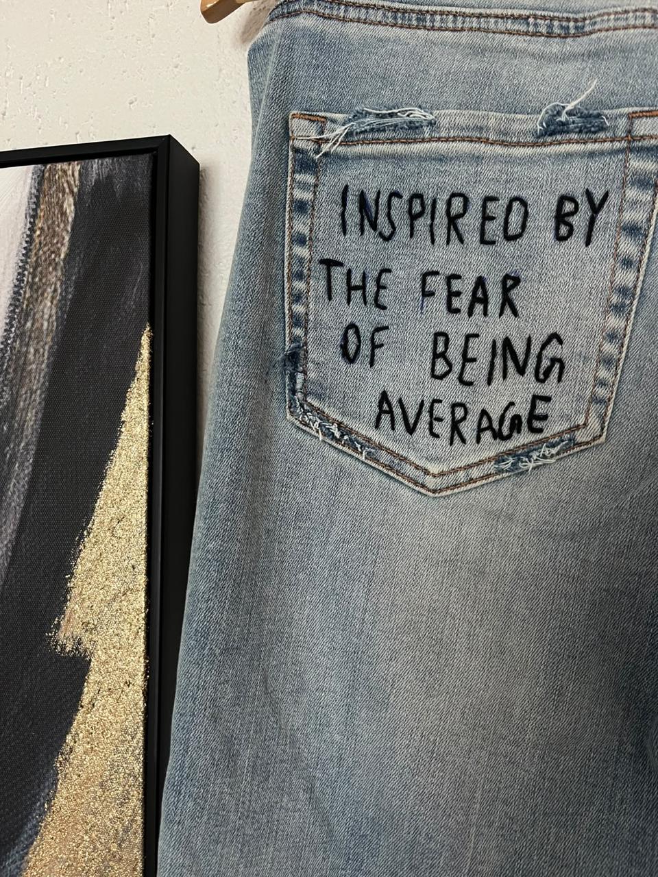 Close up of a jeans back pocket hand embroidered with 'Inspired by the fear of being average'