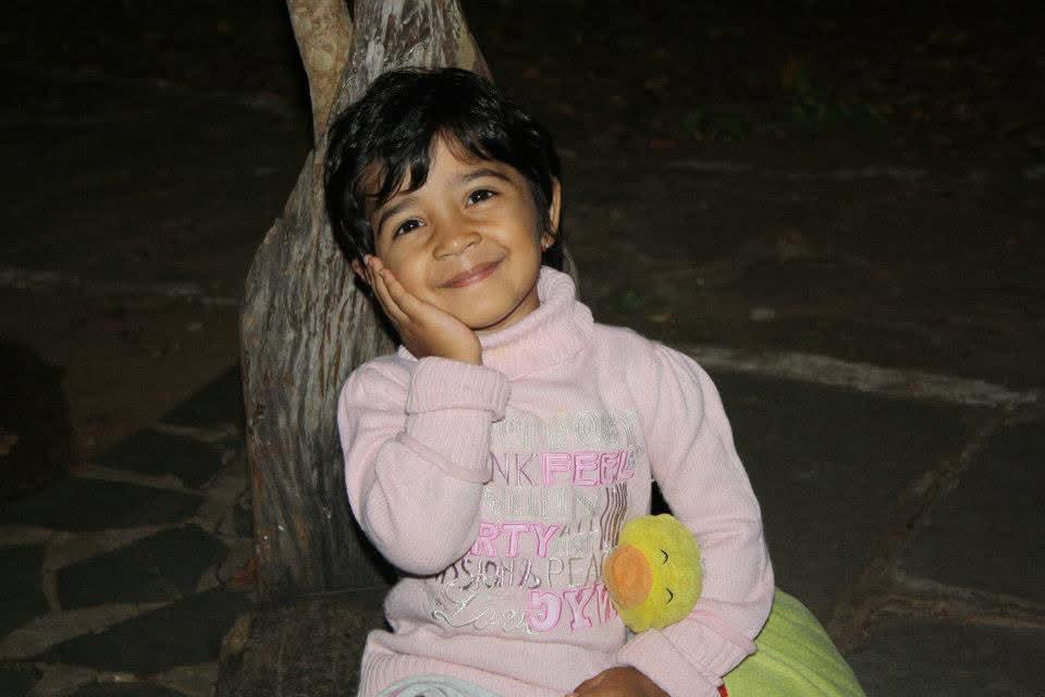 Anika as a little girl in a pink jumper, cheek resting on her hand, grinning